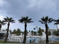 A swimming pool surrounded by sun loungers and parasols, under which people hide from the rain, surrounded by palm trees against a