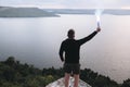 Protest concept. Brutal man holding blue flare torch in hand, standing on top of hill in evening with view on river. Ultras