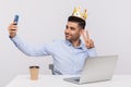 Proud happy businessman sitting in office workplace, wearing crown on head and taking selfie on mobile phone