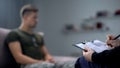 Psychologist making notes during therapy session with sad male soldier, PTSD