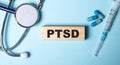 PTSD post-traumatic stress disorder is written on a wooden block on a blue background near a stethoscope, syringe and pills.