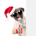 Pug puppy with retro microphone in red christmas hat peeking from behind empty board and showing thumbs up. isolated on white