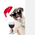 Pug puppy with wineglass and retro microphone in red christmas hat peeking from behind empty board. isolated on white background Royalty Free Stock Photo