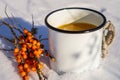 Punch with sea buckthorn berries in an iron white mug in the snow. Sea buckthorn twigs with tea