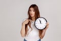 Punctual woman pointing finger at white display of big wall clock seriously looking at camera, Royalty Free Stock Photo