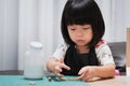 Pupil girl doing paper craft at homeschool. Kindergarten student gluing. Happy child wearing black apron. Baby aged 4-5 years old