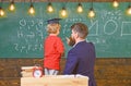 Pupil in graduation cap solving equation with the help of the teacher. Man teaching little kid arithmetic while pointing Royalty Free Stock Photo