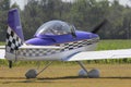 A Purple, Black, and white Airplane at a Flying Farmers show sitting by the runway by the airplane hanger on a colorful day.