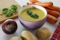 Purple bowl of potato and carrot cream soup with ingredients, wooden cutting board and red kitchen towel on white table Royalty Free Stock Photo