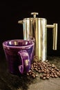 Purple cup and chrome French press and coffee beans against black background sitting of rough slate surface