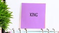 A purple notepad with the word KING written on it is attached to a notebook with a houseplant beside it. The notepad is square and Royalty Free Stock Photo