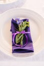 A purple serving napkin with a sprig of green leaves and a cornflower, tied with a satin ribbon, lies on a serving plate
