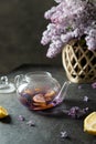 Purple tea with lemon in a transparent teapot and a bouquet of blooming lilac on a dark background. Spring tea drinking. Lilac tea