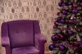 Purple velvet chair near artificial Christmas tree with purple Christmas balls and violet tinsel garlands at wallpaper background. Royalty Free Stock Photo