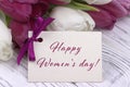 Purple and white tulips with white paper on a white wooden background and card lettering happy women`s day english. Womans Day. 8 Royalty Free Stock Photo