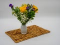 Purple, Yellow and Green floral arrangement in a white glass vase sitting on a brown whicker placemat with a white background