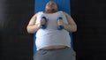Purposeful fat man lifting dumbbells with great effort lying on mat, top view