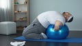 Purposeful overweight male lifting dumbbells lying on fitness ball, muscles pump