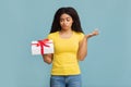 Puzzled african american woman holding gift decorated with ribbon, standing over blue background, studio shot Royalty Free Stock Photo