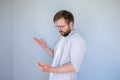 Puzzled bearded man in glasses and casual clothes looks at smartphone, he is confused and makes a hand gesture. Gray Royalty Free Stock Photo