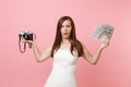 Puzzled bride woman in wedding dress holding retro vintage photo camera, bundle lots of dollars, cash money choosing