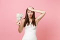 Puzzled bride woman in white wedding dress keeping hand on forehead holding one dollar bills on pastel pink