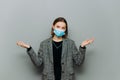 Puzzled business woman in suit and medical mask on face isolated on gray background, spreads her arms to the sides