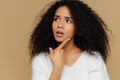 Puzzled female model keeps fore finger on chin, looks thoughtfully aside, has frustrated facial expression, curly Afro hair, wears