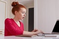 Puzzled female talented editor of online issue works remotely at home, dressed casually, has red hair tied in hair bun, creats art