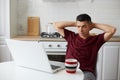 Puzzled freelancer man wearing casual style clothing sitting in front of portable computer in kitchen, keeps arms behind head, Royalty Free Stock Photo