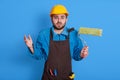 Puzzled repairman wears hardhat, apron, and uniform, holds paint roller in hand for painting wall, craftsman uses special tool,