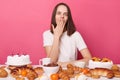 Puzzled surprised brown haired woman in white t shirt sitting at table with lots desserts breaking diet covering mouth with palm