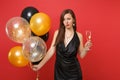 Puzzled upset girl in little black dress celebrating holding glass of champagne air balloons isolated on red background