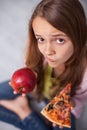 Puzzled young girl undecided wheter to choose the appetizing pizza or healthy apple Royalty Free Stock Photo