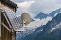 PV Solar Panels And Satellite Dish Antenna At The Wall Of A Wooden Building With Snowy Mountains In The Background