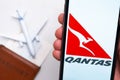 Qantas Airline application on the screen of mobile phone in mans hand. Passport, boarding bass are next to a white plane