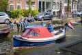 Quaint small blue boat with red canopy carrying Dutch flag cruising on canal past group young adults sitting on wall