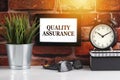 QUALITY ASSURANCE text with alarm clock, books and vase on brick background Royalty Free Stock Photo