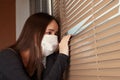 Quarantine and self-isolation. A young girl in an antibacterial mask looks out the window from the house to the street, sits in