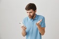 Questioned good-looking guy looking at screen of smartphone, lifting hand cause he is clueless and annoyed with what he