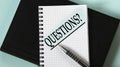 QUESTIONS? - word in a white notebook against the background of a black notebook with a pen