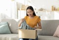 Quick purchases in online store. Satisfied asian woman opening carton box sitting on sofa in living room at home