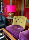 Eclectic living room with brightly coloured velvet upholstered chairs, pink lampshade and deep red patterned carpet.