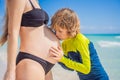 A radiant pregnant mother and her excited son share a tender moment on a serene, snow-white beach, celebrating