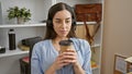 Radiant young, hispanic woman basks in her early morning office success, confidently sipping coffee while immersed in her work, Royalty Free Stock Photo