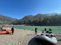 a raft and several people standing near a river with mountains in the background Royalty Free Stock Photo