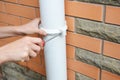 Rain gutter installation: a man is screwing the downpipe bracket, socket clip with a screwdriver to fix the downspout to the wall