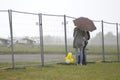 In the rain. Man and woman standing under the umbrella behind the fence, landing field with planes on the background