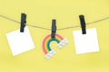 Rainbow figurine and blank note sheets attached to rope with black wooden clothespins against yellow background, copy space.