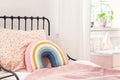 Rainbow pillow and pink sheets on girl`s bed in bright bedroom interior with plant.